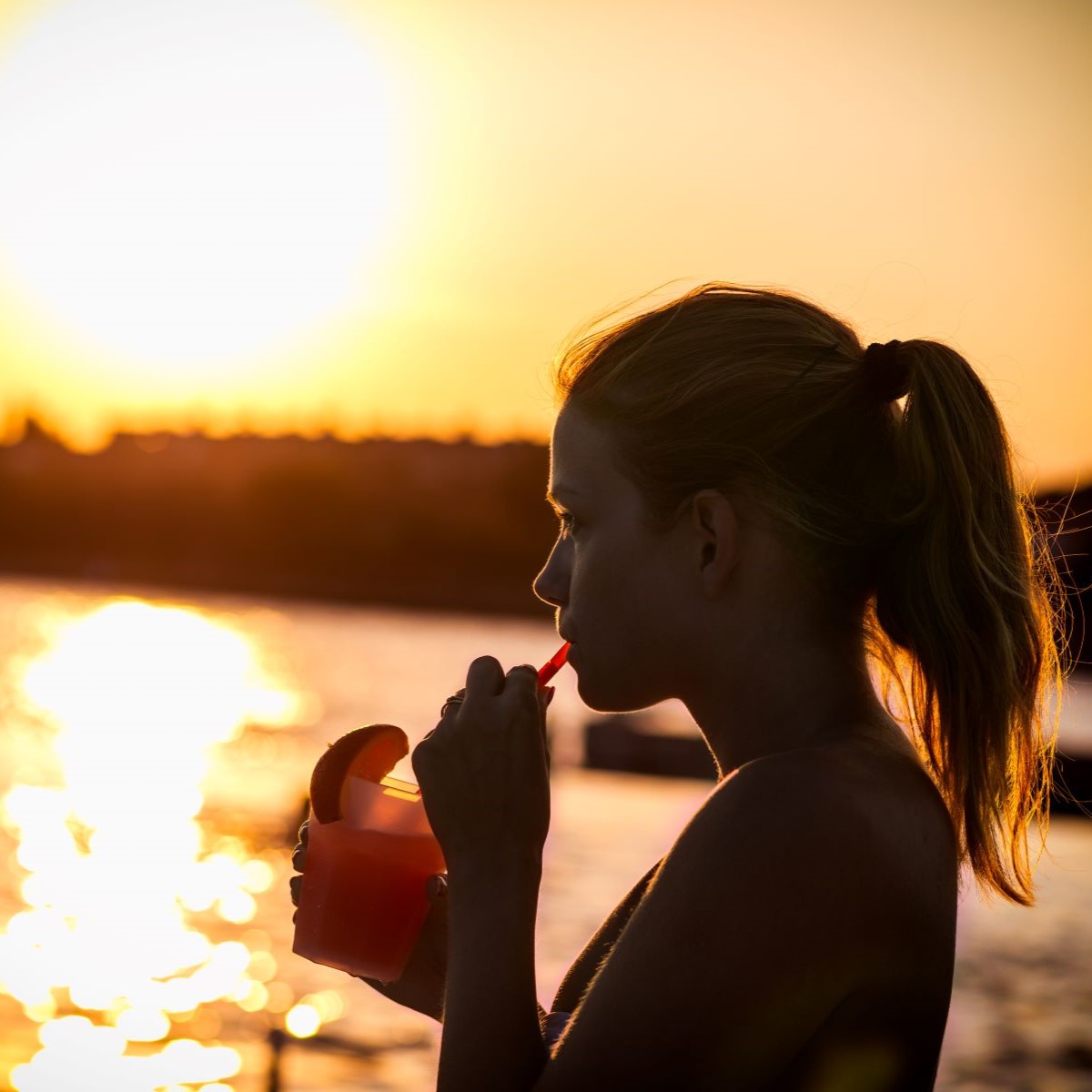 Frau mit Zopf trinkt einen Cocktail im Sonnenuntergang